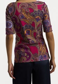 Mujer vestida con una blusa ajustada magenta de mangas cortas, que presenta intrincados patrones paisley en púrpura, dorado y azul, combinada con pantalones oscuros.