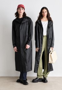 Sort vandtætte lange frakker med hætte, frontlommer og trykknaplukninger. Én model har grønne bukser på; den anden har mørke jeans og en kasket.
