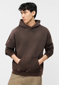 Νέος άνδρας που φοράει καφέ φούτερ με κουκούλα, με τα χέρια στην μπροστινή τσέπη, κοιτάζοντας προς το πλάι σε απλό λευκό φόντο.