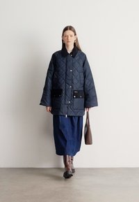 Tengerészkék steppelt dzseki magas gallérral, fekete részletekkel ellátott zsebekkel, gombos rögzítéssel, kék szoknya és barna csizma fölött viselve, barna táskával tartva.