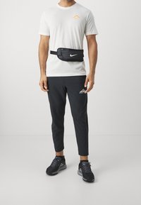 Riñonera negra con el logo blanco de Nike, llevada sobre una camiseta blanca y pantalones deportivos negros, complementada con zapatillas de running negras.