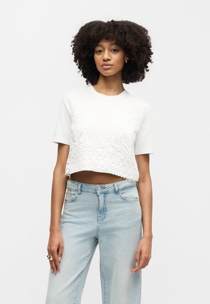 Kvinde med naturligt krøllet hår iført en hvid blonde crop top og lyseblå jeans med høj talje, stående foran en ensfarvet baggrund.