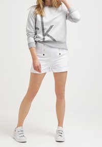 Sudadera gris con un logo circular, combinada con pantalones cortos blancos de talle alto que presentan botones dorados en la parte delantera. El look se completa con zapatillas plateadas.