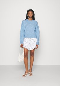 Suéter azul claro, saia curta branca com padrão floral e sandálias transparentes com tiras. A segurar uma bolsa de palha branca.