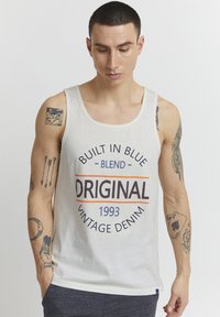 Top din bumbac alb, cu textul "Built in Blue," "Original," și "Vintage Denim 1993" în albastru și portocaliu. Modelul afișează tatuaje pe brațe.