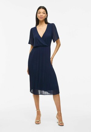 Femme portant une robe midi plissée bleu marine à manches courtes et décolleté en V profond, associée à des sandales à talons hauts dorées, se tenant sur fond blanc.
