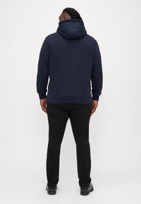 Sötétkék kapucnis pulóver hosszú ujjal, kenguruzsebbel és bordás mandzsettával. Feketementes farmernadrággal és fekete cipővel párosítva.