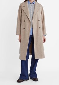 Beżowy płaszcz podwójnie zapięty z wełny z dużymi klapami, zestawiony z jasnoszarym swetrem w warkocze i niebieską koszulą w paski, noszony z dzwonami.
