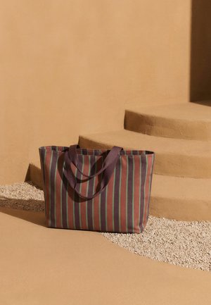 Braune Tragetasche mit grünen und roten vertikalen Streifen, die auf sandigem Boden nahe geschwungener beiger Stufen liegt.