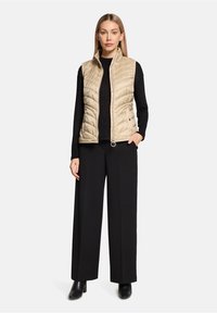 Veste matelassée beige avec col montant, fermeture éclair et deux poches latérales, portée sur un pull noir ajusté et un pantalon noir à jambes larges.