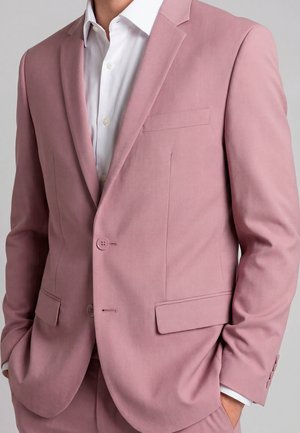 Man draagt een op maat gemaakte stofroze colbert met bijpassende broek en een wit overhemd, staand met handen in de zakken.