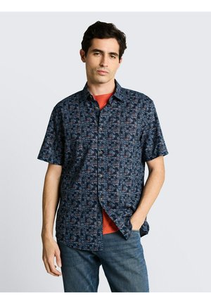 Mężczyzna w granatowej, krótkiej koszuli zapinanej na guziki z geometrycznym wzorem, na rdzawym T-shircie i niebieskich dżinsach, stojący z jedną ręką w kieszeni.