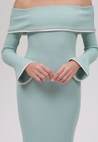 Mintgrønn off-the-shoulder kjole med teksturert stoff, kontrasterende hvit kant på ermene og flagrende mansjetter. Figurtilpasset silhuett.