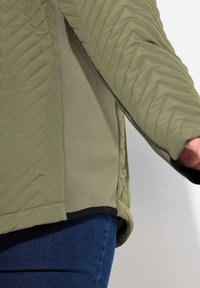 Olijfgroene jas met een doorgestikt chevronpatroon, zachte textuur en zwarte rand aan de onderkant. Close-up toont details van de arm en zijkant.
