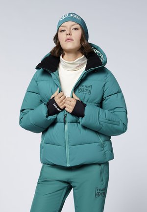 Young woman wearing teal winter jacket, matching pants, and beanie, holding jacket zipper near collar, with neutral expression.