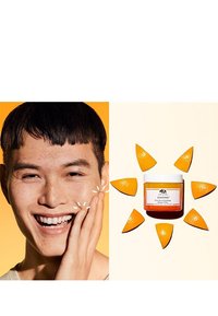 Un pot orange de masque éclaircissant GinZing avec un couvercle blanc, entouré de tranches de pelure d'orange sur un fond pêche pâle. Une personne souriante touchant son visage.