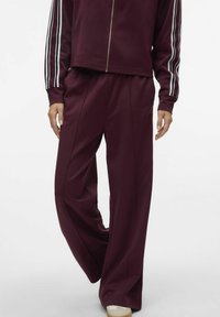 Mörkröd sportoutfit bestående av vida byxor med sidosömmar och en matchande zip-up tröja med vita randiga detaljer på ärmarna.