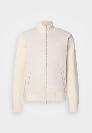 Helle beige Jacke mit durchgehendem Reißverschluss, bestehend aus glattem Obermaterial und strukturierten Strickärmeln, ausgestattet mit seitlichen Taschen und einem Stehkragen.