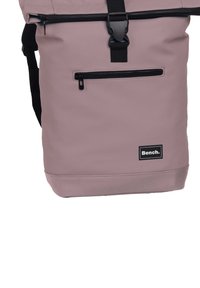 Roze roll-top rugzak met een zwarte rits, verstelbare schouderbanden en een voorzak met rits. Bevat een zwart logo label op de onderste voorkant.