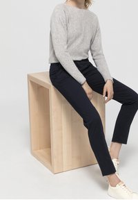 Grå, långärmad tröja i kombination med marinblå chinos; sitter på en träkub. Vita sneakers med beige snören. Enkel, avslappnad outfit.