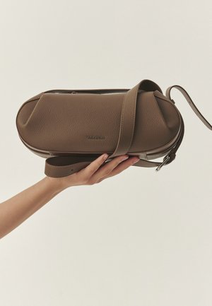 Mână ținând o geantă maro din piele, tip crossbody, cu o curea și o cataramă metalică, având un mic nume de brand embossat pe partea din față.