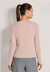 Frau mit einem dunklen Pferdeschwanz, die einen hellrosa gerippten Pullover mit gewellten Kanten und grauen Hosen trägt, mit dem Rücken zu einer schlichten Hintergrund.