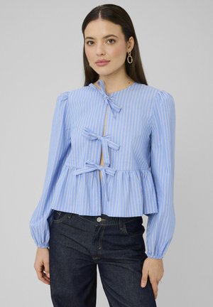 Femme portant un chemisier à manches longues rayé bleu et blanc avec trois nœuds à l'avant et un ourlet évasé, associé à un jean bleu foncé.