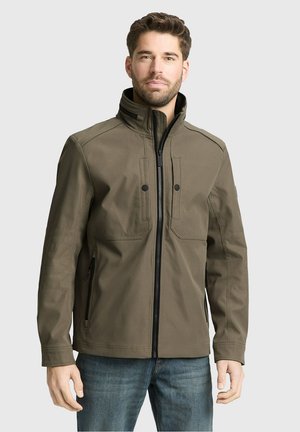 Mann trägt eine olivgrüne Jacke mit Reißverschluss und hohem Kragen sowie blaue Jeans, steht vor einem schlichten hellen Hintergrund.