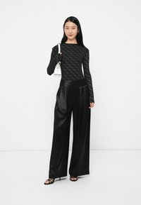 Donna che indossa un top a maniche lunghe nero con un motivo, pantaloni larghi in satin neri a vita alta, sandali con tacco neri e tiene in mano una piccola borsa a spalla bianca.