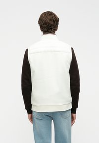 Vyras su garbanotais rudais plaukais, vilkintis baltą užpildytą liemenę ant juodos ilgomis rankovėmis marškinėlių ir šviesiai mėlynų džinsų, atsisukęs nugara prieš baltą foną.