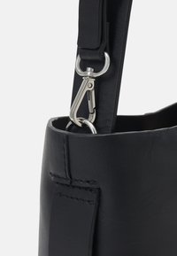 Détail d'un sac en cuir noir mettant en avant le matériau texturé, une pince argentée et des accents de couture sur la sangle et l'ouverture du sac.