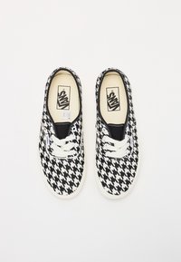 Kankaan kengät, joissa on mustavalkoinen houndstooth-kuosi, valkoiset nauhat ja kumipohja. Kengissä on musta vuori ja logo pohjallisessa.