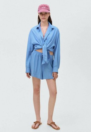 Lichtblauw cropped shirt met een vastgebonden voorkant, bijpassende shorts en een roze baseballcap met de tekst "NEW YORK"; eenvoudige bruine flip-flops.