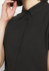 Chemise noire à col en coton ; présente un devant à boutons, des manches courtes et une texture lisse avec un détail de couture subtil.