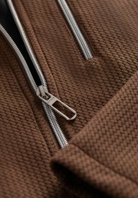 Hnědá texturovaná tkanina s diagonálním vzorem; má stříbrný zip a táhlo. Zaměření na detail zipové oblasti a materiálu.