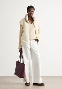 Gilet in pile beige con colletto alto, top beige chiaro, pantaloni bianchi a gamba larga e borsa tote bordeaux. Scarpe marroni con suola piatta.