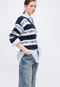 Γυναίκα με αλογοουρά που φοράει μπλε-σκούρο ριγέ πουλόβερ Tommy Jeans πάνω από ριγέ λευκό πουκάμισο και ανοιχτό μπλε σκισμένα τζιν, με τα χέρια στις τσέπες.