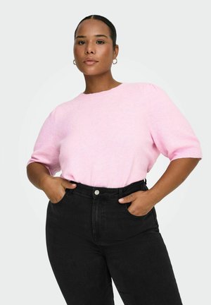 Femeie purtând un pulover roz deschis cu mâneci scurte și blugi negri cu talie înaltă, cu mâinile în buzunare, pe un fundal deschis și simplu.