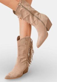 Bottes chelsea en suède tan avec des franges, bout pointu et talon empilé. Fermeture zippée sur le côté, texture lisse dans l'ensemble.