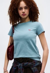 Νεαρή γυναίκα με σγουρά μαλλιά, που φοράει ένα ανοιχτό μπλε T-shirt της Levi’s, σκούρο παντελόνι, πουκάμισο καρό δεμένο στη μέση και κρατάει μια κόκκινη τσάντα ώμου.