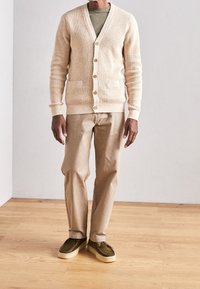 Beige cardigan with button closure and two front pockets over a green shirt, paired with light beige pants and olive green shoes.