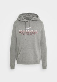 Grå huvtröja med kenguruficka, med en vit och röd "HOLLISTER CALIFORNIA"-logotyp. Tillverkad av mjukt tyg med ribbade muddar.