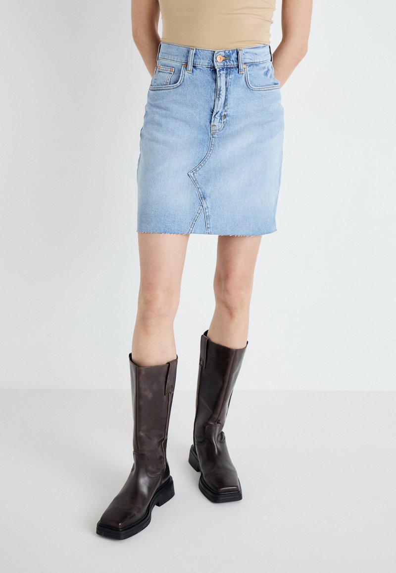 Light blue denim mini skirt with a frayed hem, featuring a front pocket and zipper closure. Paired with knee-high, shiny brown leather boots.