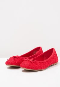 Un paio di ballerine rosse in pizzo con piccoli fiocchi sulle punte e suole piatte color sabbia, esposte su una superficie bianca con uno sfondo semplice.