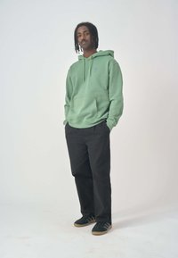 Green hoodie with a front pocket, drawstring, and small logo. Paired with black pants and black shoes, displayed against a white background.