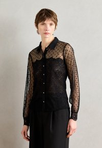 Blouse noire transparente avec motif à pois et détails en dentelle, dotée d'un col, d'une fermeture à boutons à l'avant et de manches longues avec poignets en dentelle.