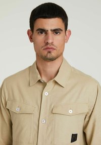Beige Hemd mit Knopfleiste aus einem glatten Stoff, ausgestattet mit zwei Brusttaschen mit weißen Knöpfen und einem kleinen schwarzen Logo.
