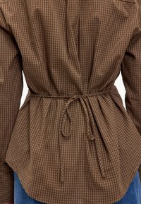 Chemise à carreaux marron avec taille cintrée et lien ajustable, dotée de manches longues et d'un ourlet arrondi. Tissu en coton texturé.
