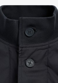 Sort jakke med krave i ribbet tekstur, to-knappet stolpelukning med runde sorte knapper og glat stof med diskret glans.