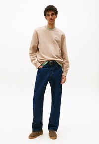 Jonge man in een beige sweatshirt, groene onderhemd, donkerblauwe jeans en bruine sneakers, staand met één hand in zijn zak.